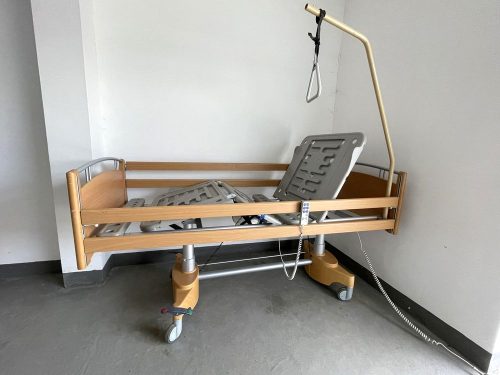Elektromos betegágy megemelt fekvőfelülettel, felhajtott oldalkorlátokkal és döntött fejtámlával, kapaszkodóval és távirányítóval.
