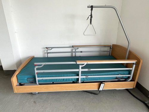 Az ágy teleszkópos leesésgátló korlátai behajtott állapotban láthatók, biztosítva a könnyű hozzáférést és a maximális biztonságot. Az állítható kapaszkodó és a kényelmes matrac további komfortot nyújtanak a használójának.