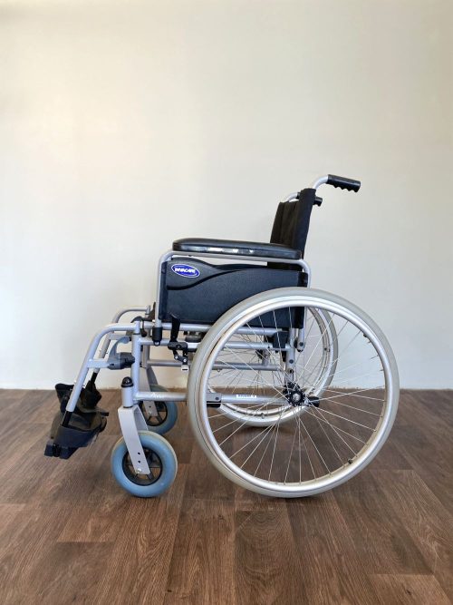 Oldalnézetből fotózott Invacare kézi kerekesszék ezüst vázzal, fekete ülőfelülettel, nagy hátsó és kisebb első kerekekkel, beltéri használatra.