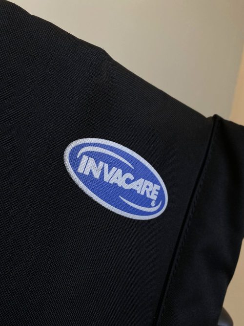 Invacare márkajelzés egy fekete szövet háttámlán, amely egy kerekesszék részét képezi.