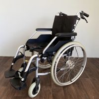 Összecsukható kézi kerekesszék masszív fémvázzal, fekete ülőfelülettel és ergonomikus karfákkal, fa padlón világos háttér előtt.