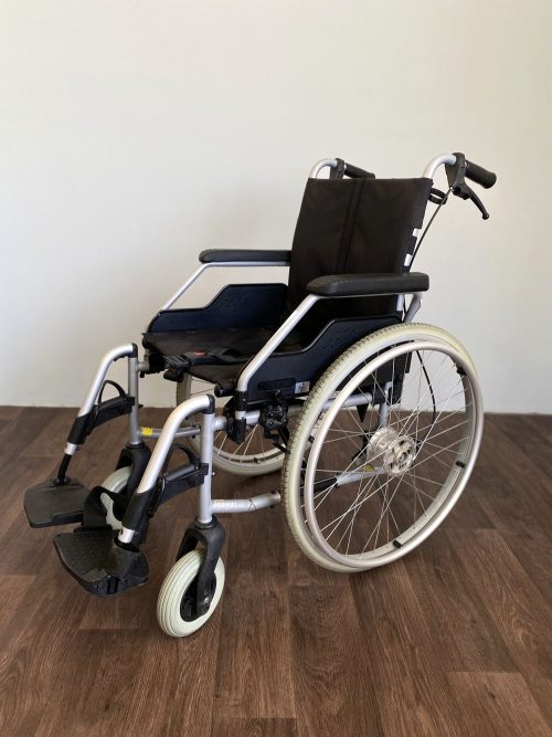 Összecsukható kézi kerekesszék masszív fémvázzal, fekete ülőfelülettel és ergonomikus karfákkal, fa padlón világos háttér előtt.