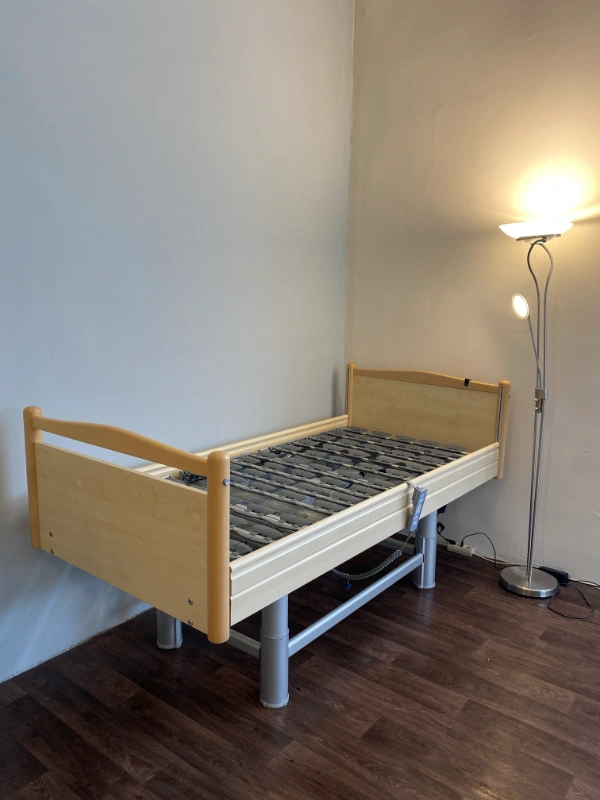 Ápolási ágy oldalnézetből, korláttal és felfüggesztett kapaszkodóval.