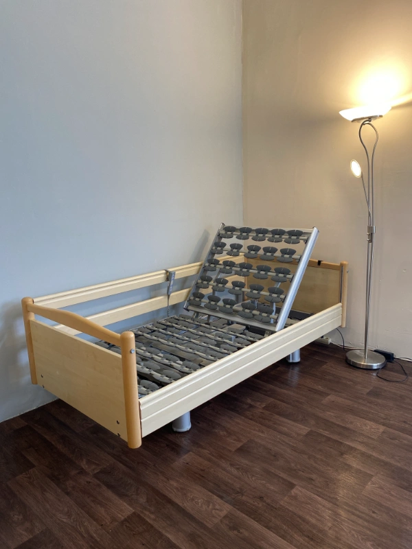 Vajszínű elektromos betegágy teljes emelésben, fal mellett elhelyezve.